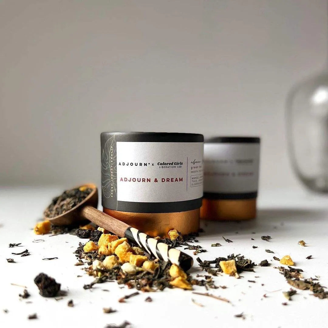 Two black-labeled cylindrical containers with copper accents on a table, surrounded by scattered herbal ingredients and a wooden scoop.